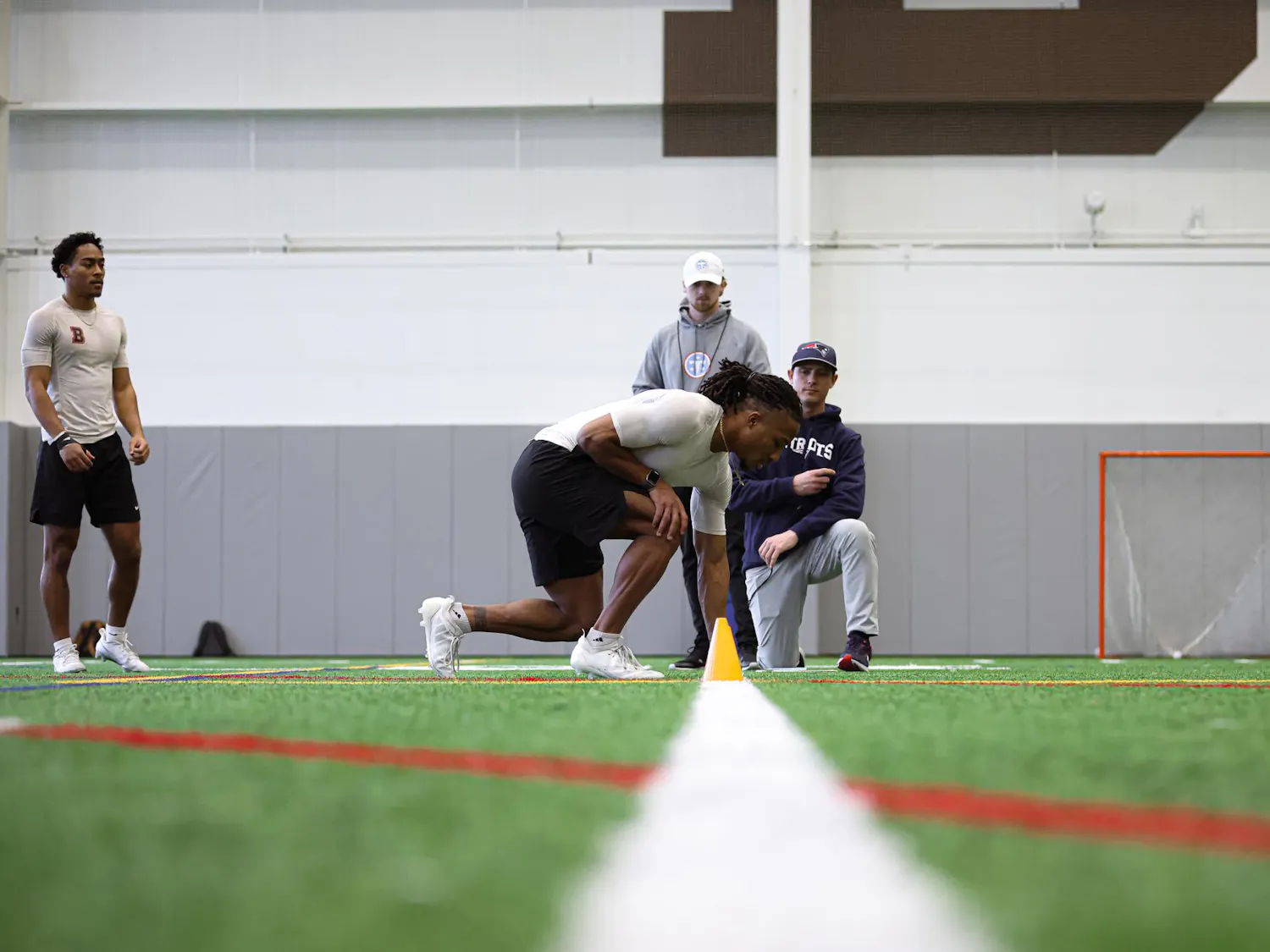 A man kneels at a start line on a turf field in front of two football scouts.