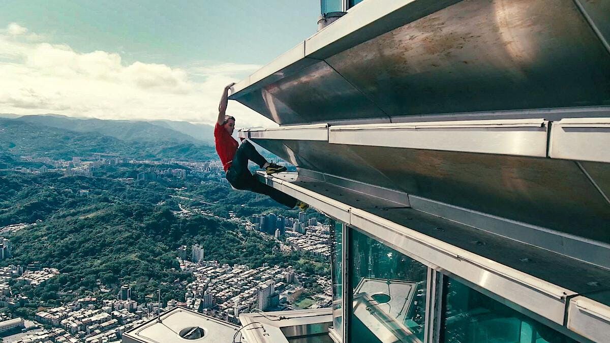 A man in a red shirt and black pants climbing on a metal building. 

