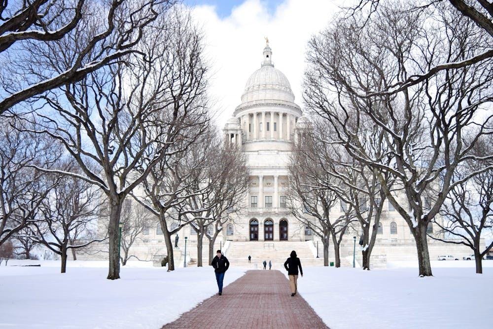 Illustration of people walking on a snow-lined pathway in front of Rhode Island's State House, a white building with a dome on top. There are also trees on either side of the path. 