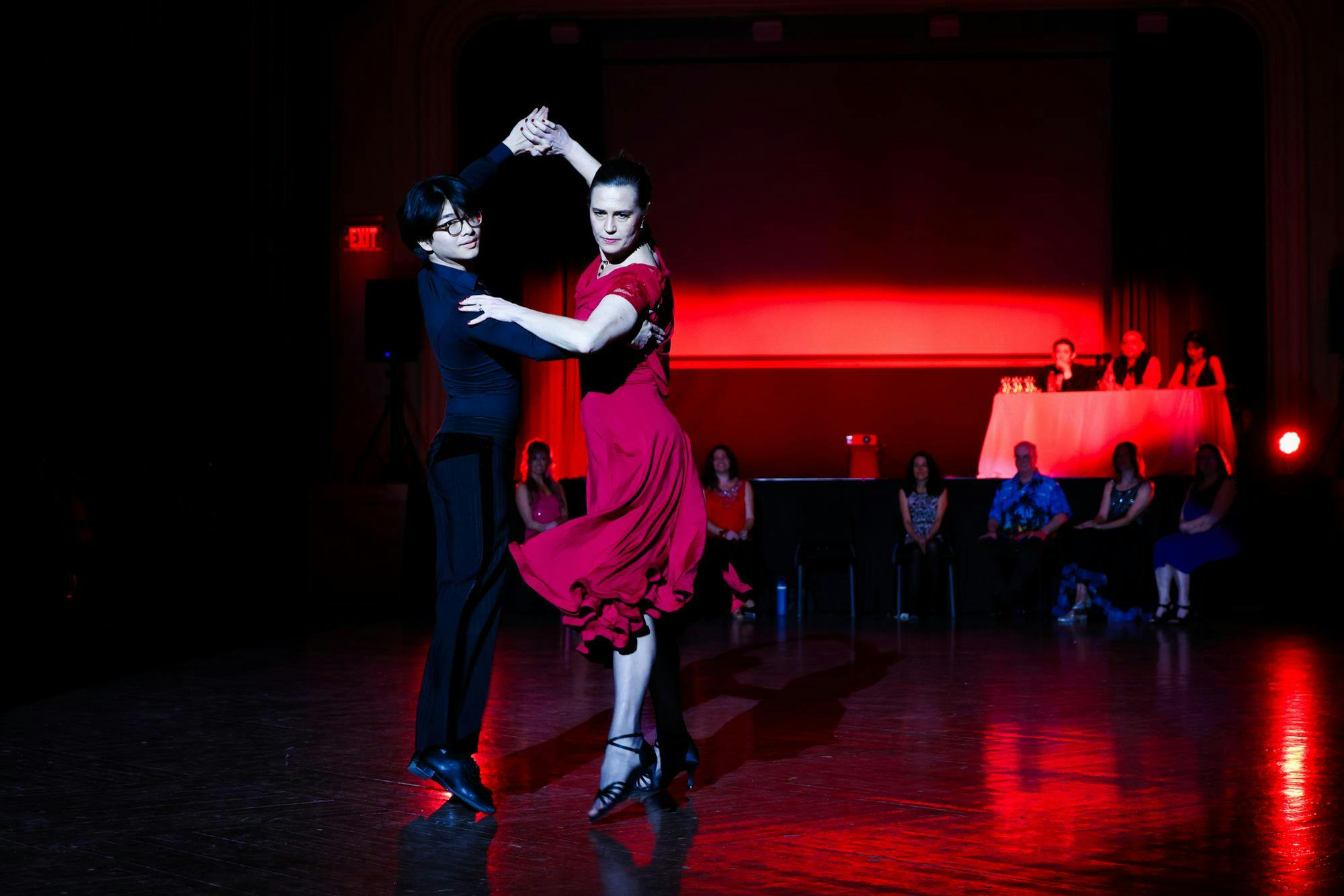 Ian Chow ‘28 and Kareen Coulombe hold a pose in their Paso Doble dance under red lights with judges and other professors looking on in the background.