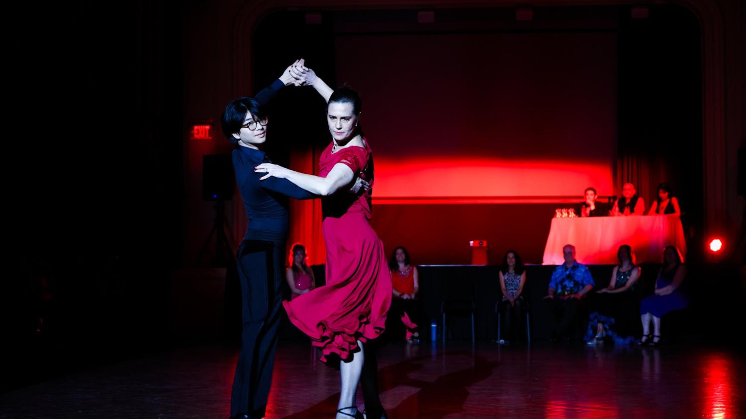 Ian Chow ‘28 and Kareen Coulombe hold a pose in their Paso Doble dance under red lights with judges and other professors looking on in the background.