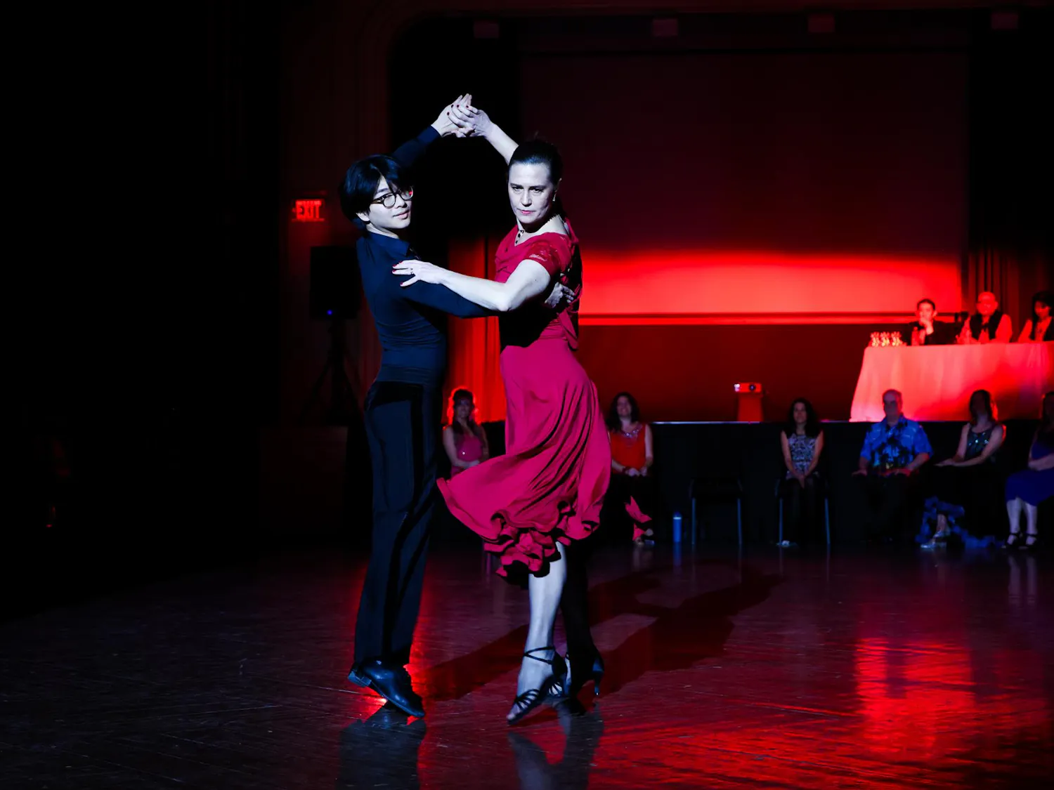 Ian Chow ‘28 and Kareen Coulombe hold a pose in their Paso Doble dance under red lights with judges and other professors looking on in the background.