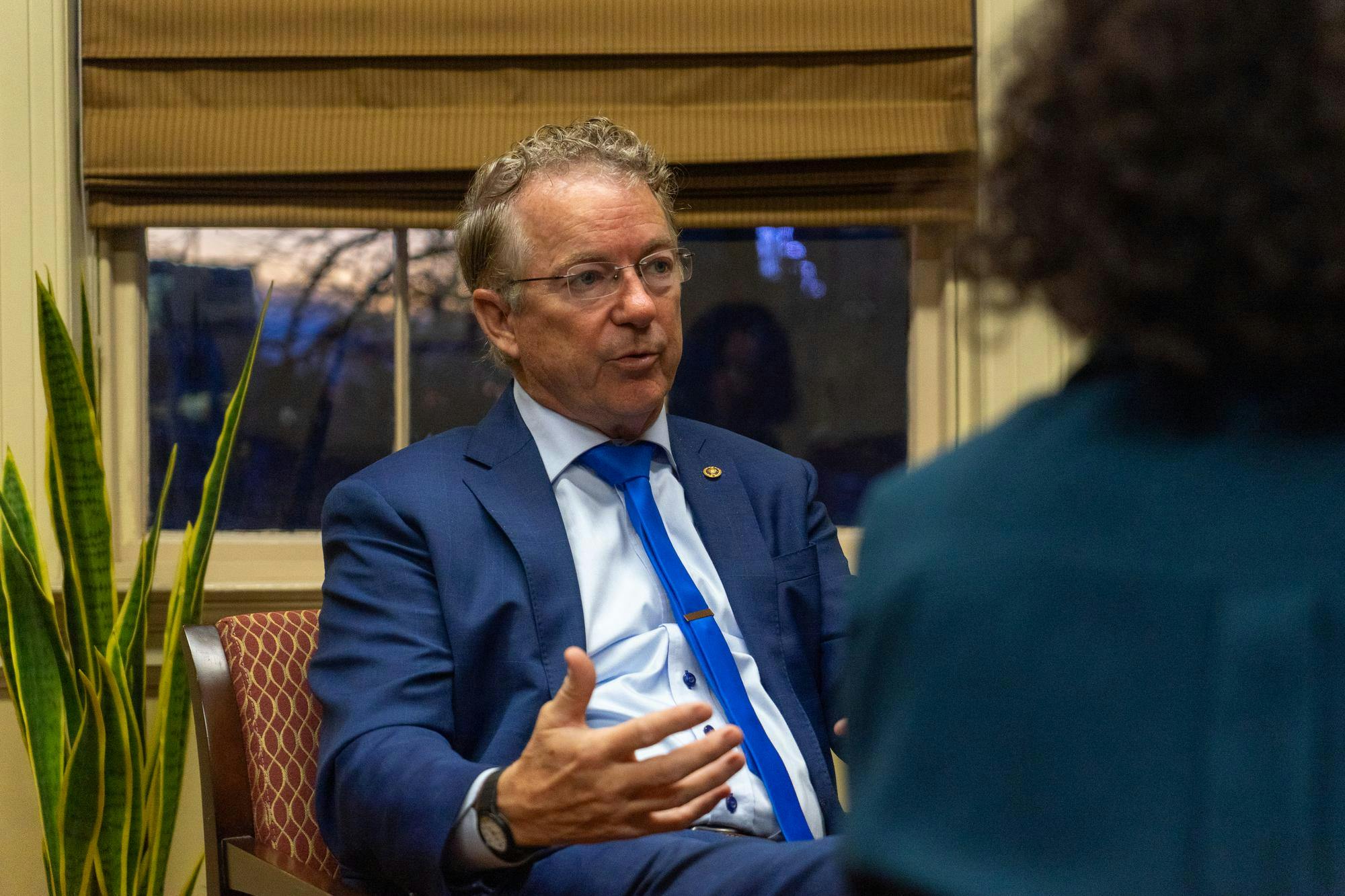 U.S. Sen. Rand Paul, a gray-haired man wearing glasses and a navy blue suit, speaks with a student while sitting in an office with a plant and a sunset in the window behind.