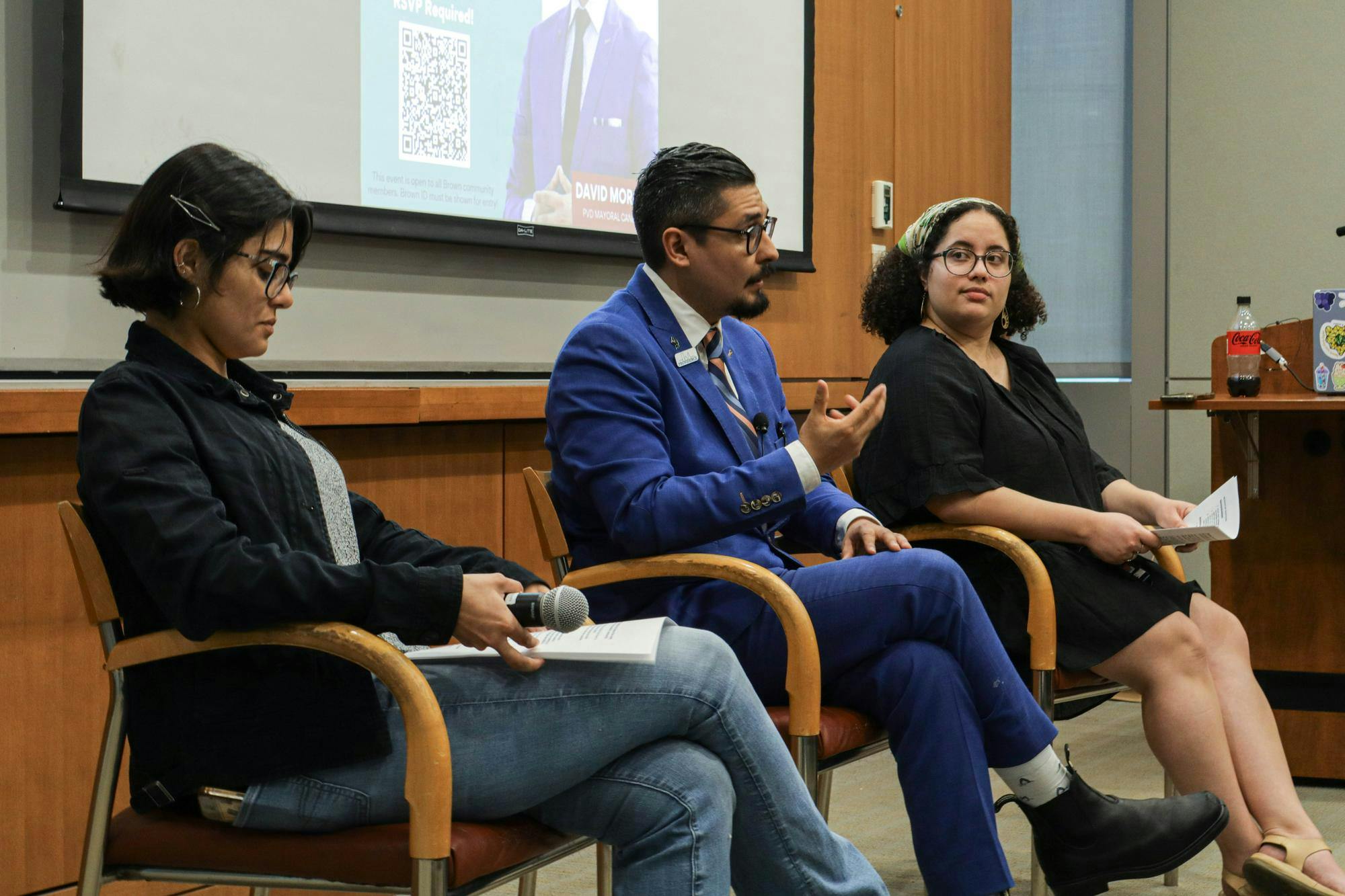 Three people sit talking in chairs in front of a presentation screen, one man and two women.