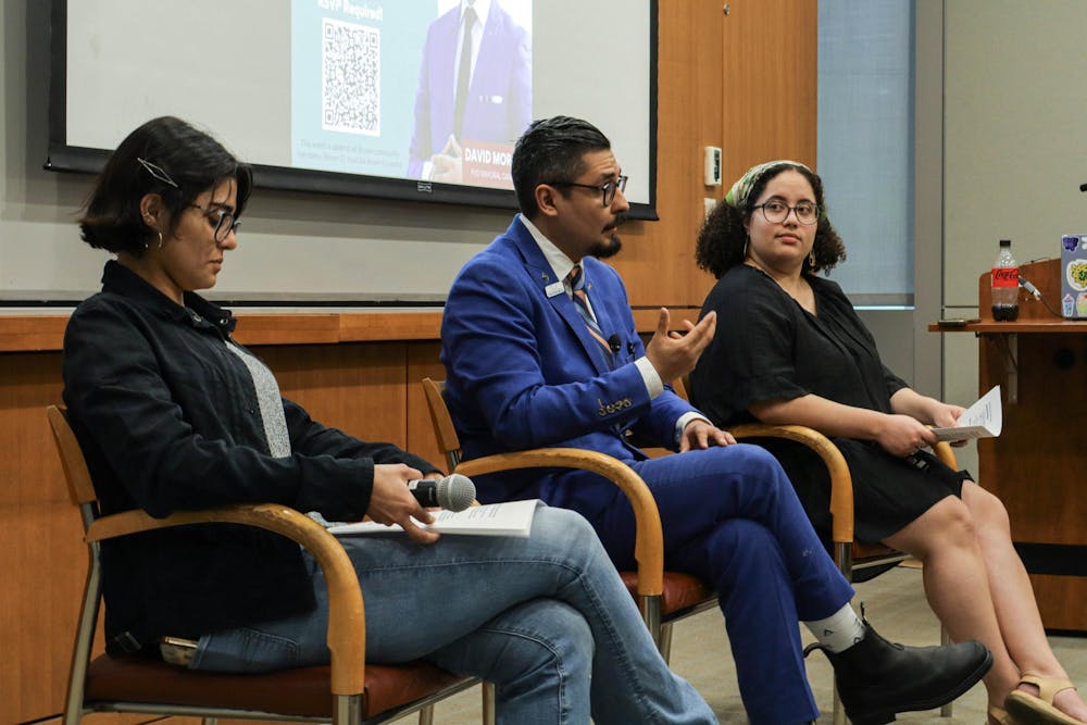 Three people sit talking in chairs in front of a presentation screen, one man and two women.