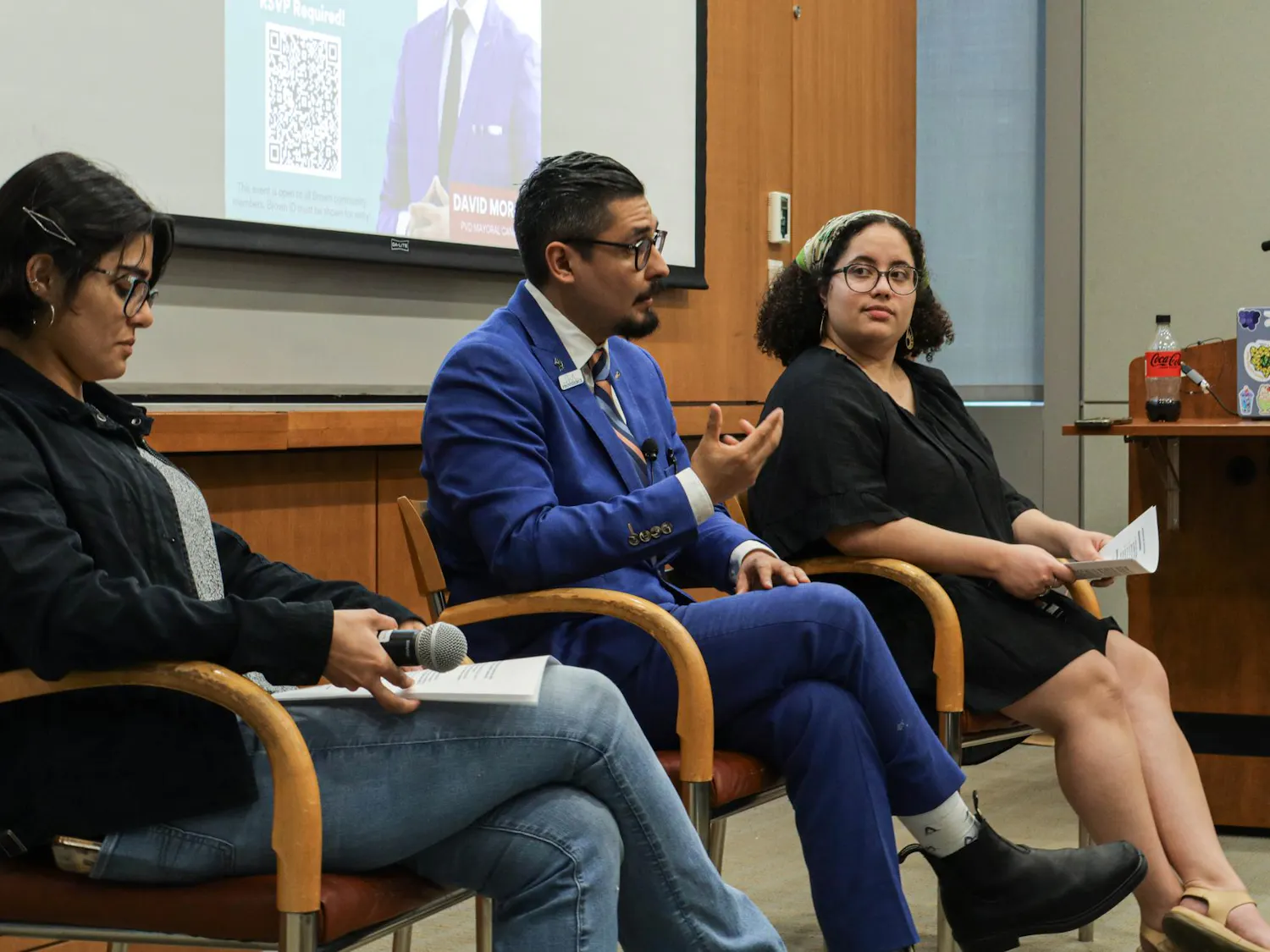 Three people sit talking in chairs in front of a presentation screen, one man and two women.