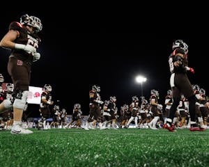 A photo taken from a low angle of many Brown football players walking onto the field. The Jumbotron and a floodlight are visible in the background.