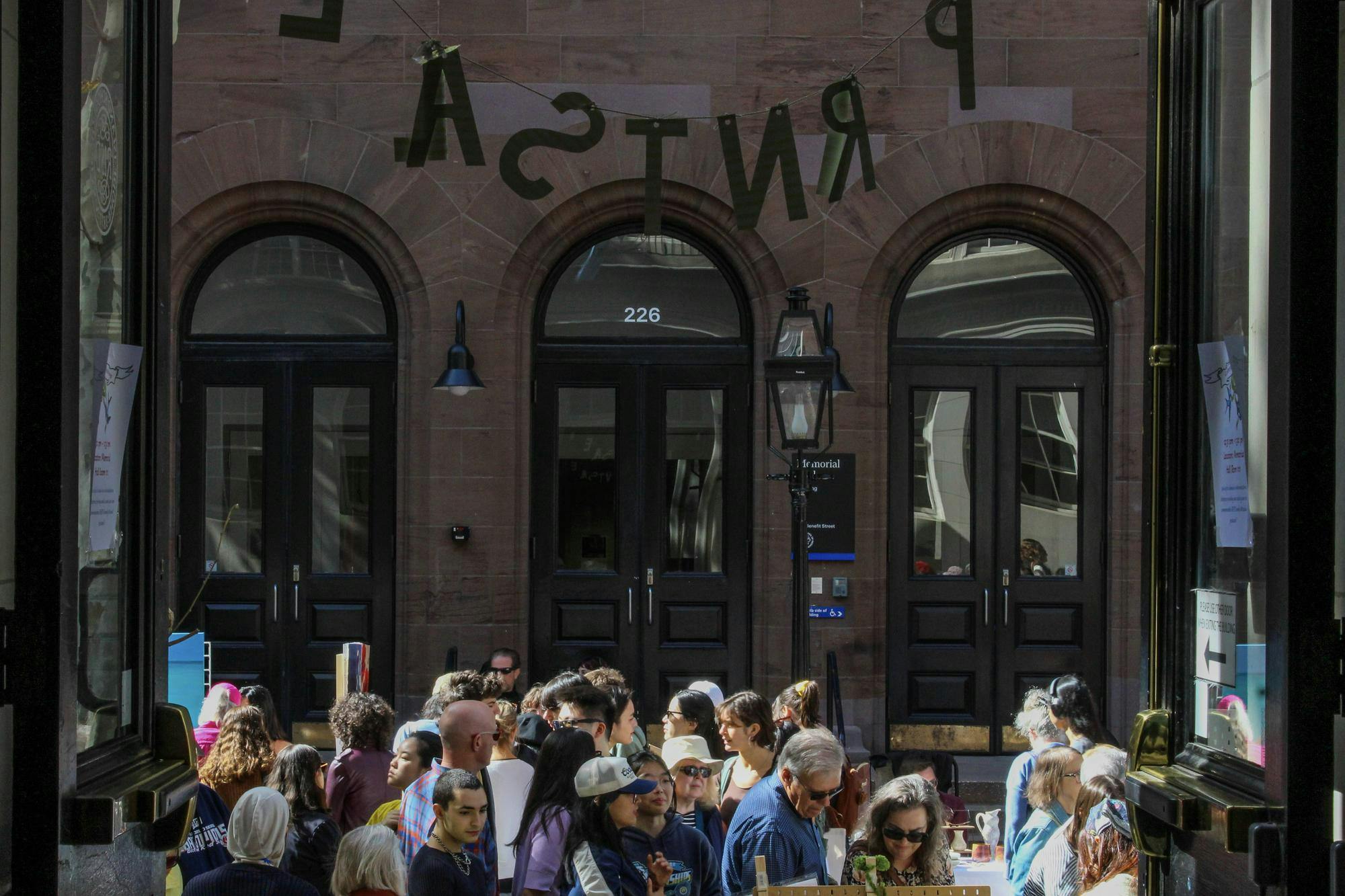 Photo of a doorway opened toward a brick building across the street with people looking at art vendors’ tables lined along the road. 