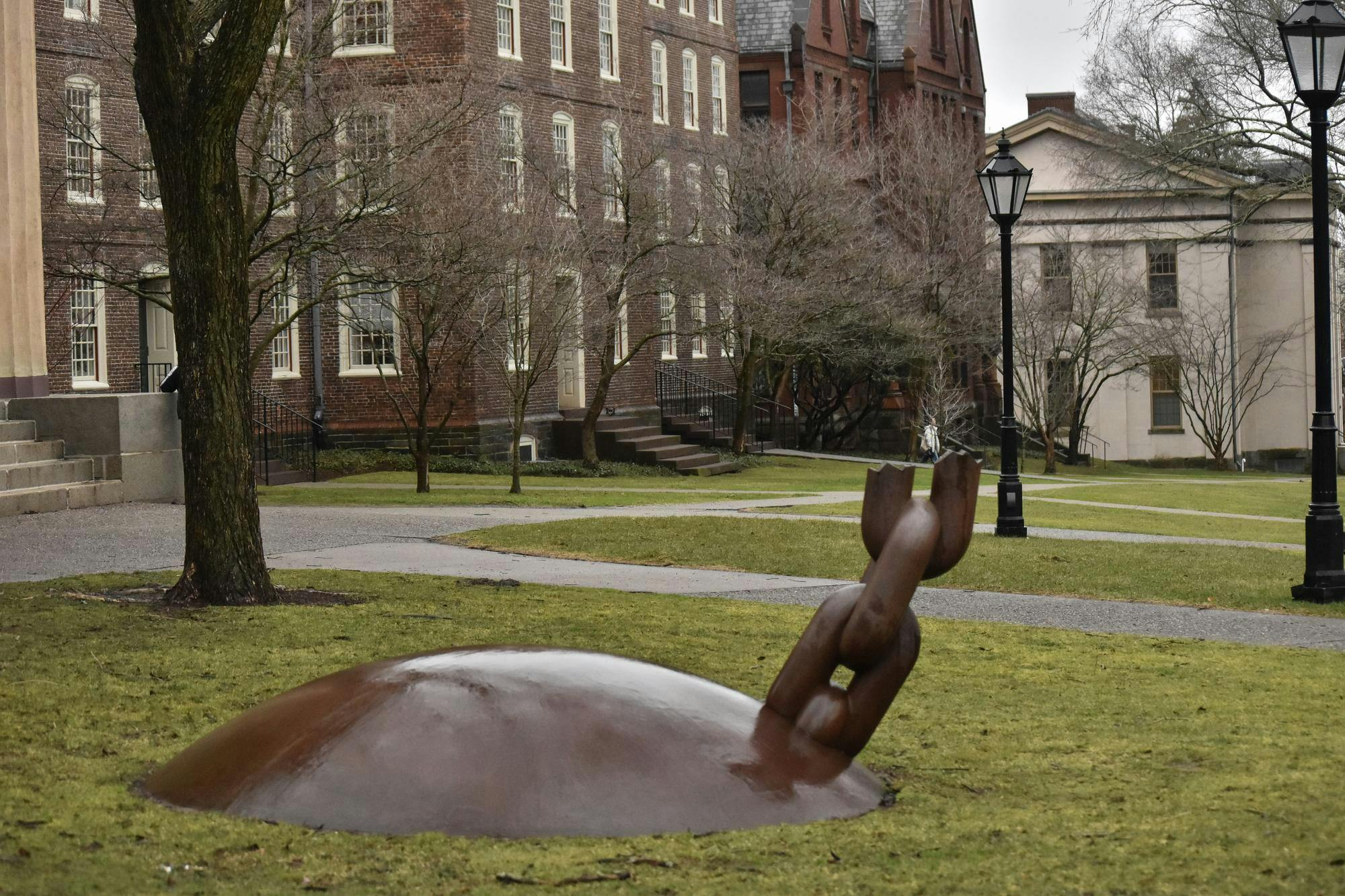 A photo showing a statue of a broken ball and chain installed in the grass of the Quiet Green with buildings, light posts and trees in the background. 