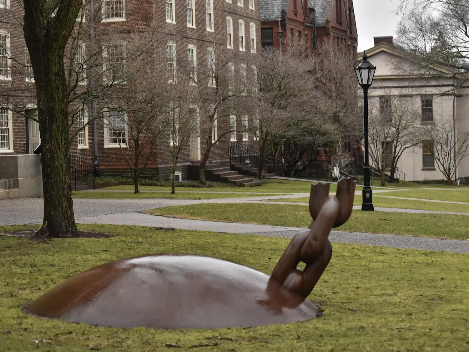A photo showing a statue of a broken ball and chain installed in the grass of the Quiet Green with buildings, light posts and trees in the background.