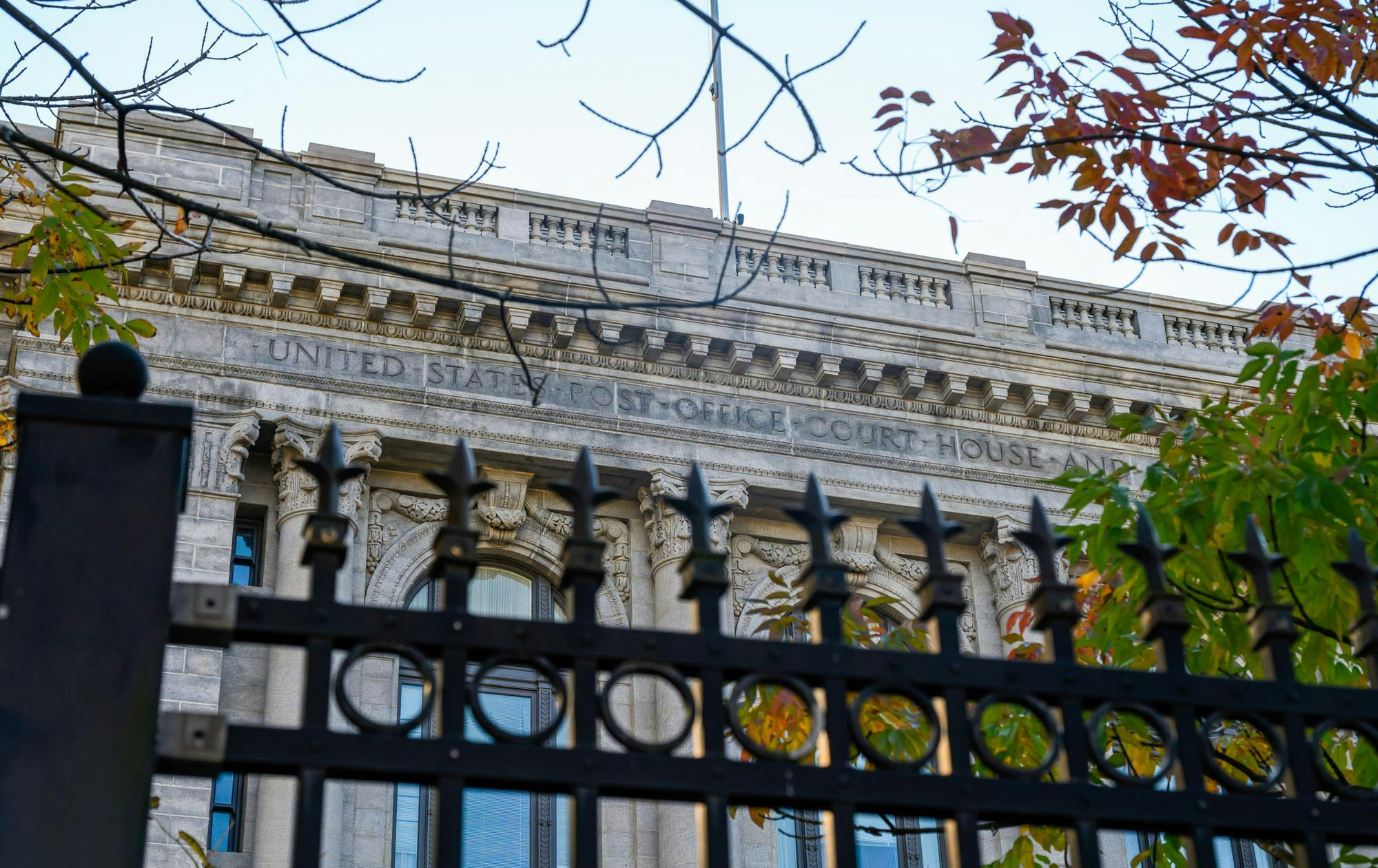 Metal gate with spikes in front. A large stone building with the words "United States Post Office Court House" etched in. A tree branch comes in from the left.
