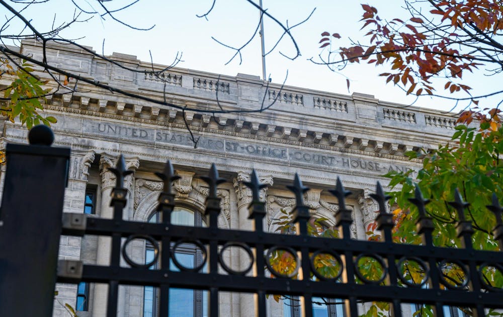Metal gate with spikes in front. A large stone building with the words "United States Post Office Court House" etched in. A tree branch comes in from the left.