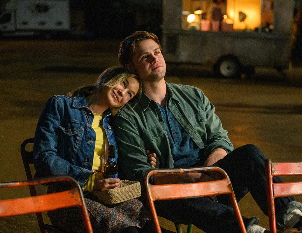 Bridget Jones (played by Renée Zellweger) rests her head on Roxster's (played by Leo Woodall) shoulder. The two are sitting outside in two chairs in a dark, nighttime setting.