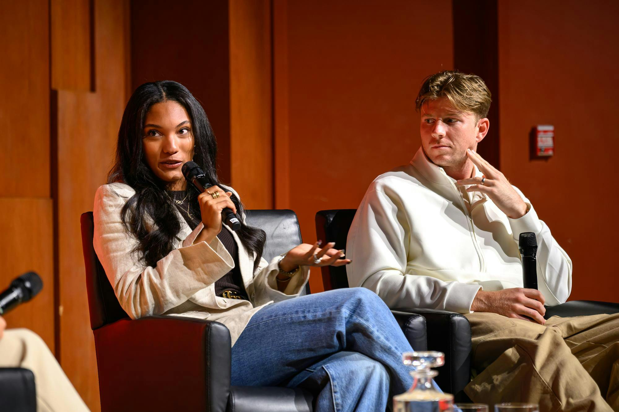 Photo of Tara Davis-Woodhall gesturing and speaking into a microphone as Hunter Woodhall looks at her. The two are seated on stage with paneled wood and a maroon wall behind them.