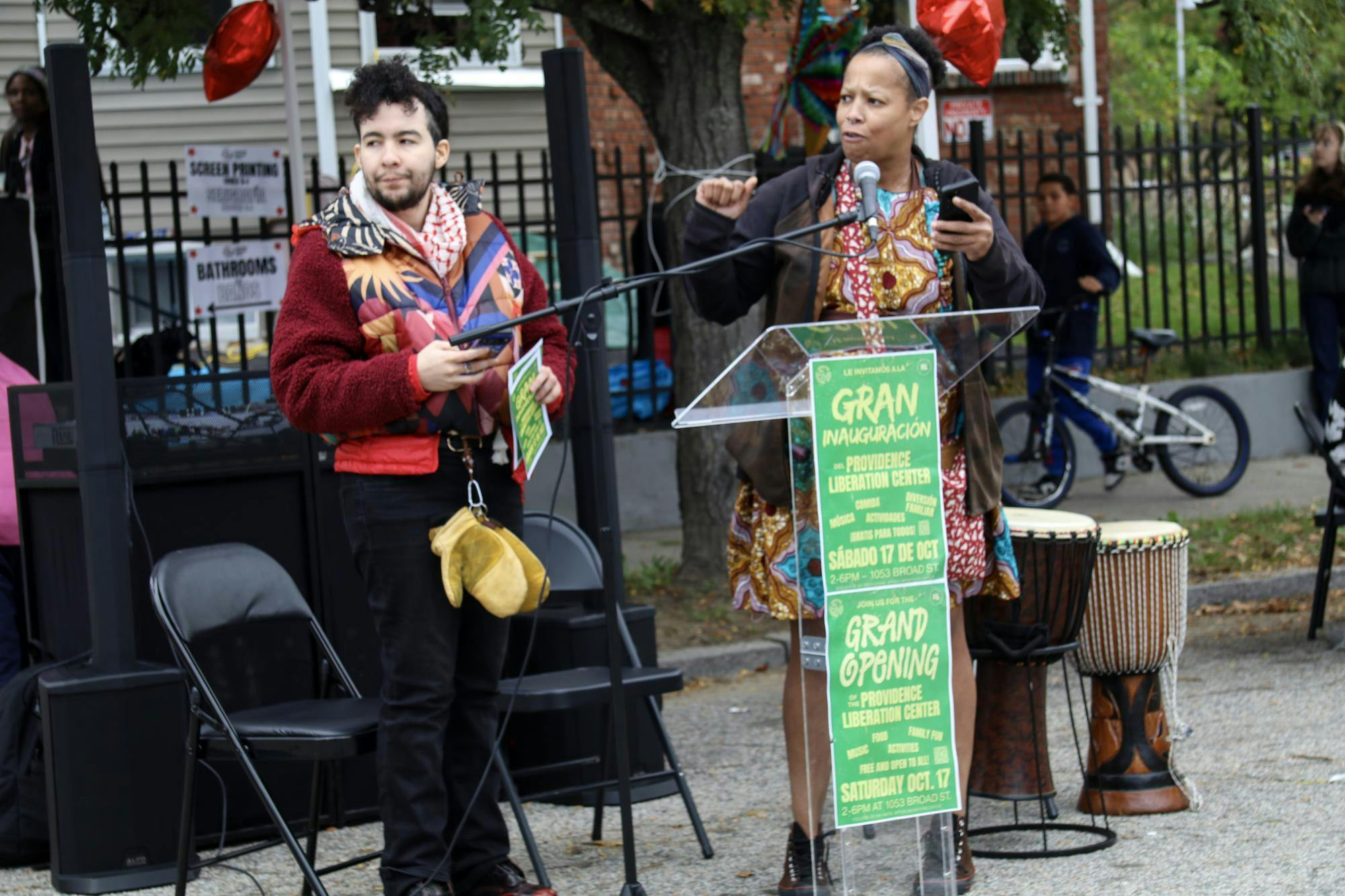 Photo of a woman wearing a colorful patterned dress speaking into a microphone and standing in front of a clear plastic lectern with a man wearing a colorful patterned vest standing on the left. 
