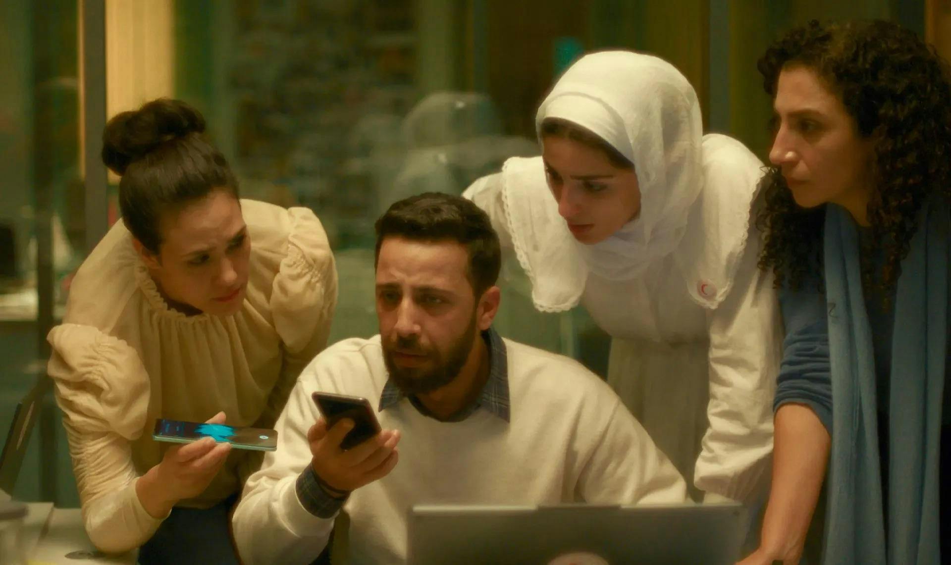 A group of three women and a man with concerned expressions on their faces listen to the phone call attentively, the woman on the left recording the conversation. 