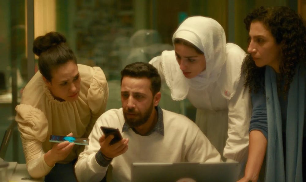 A group of three women and a man with concerned expressions on their faces listen to the phone call attentively, the woman on the left recording the conversation. 