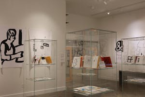 Photo of the “Everywhere I Go,” exhibit showing various clear display cases of open sketchbooks and large, bold black-and-white drawings displayed on the walls.