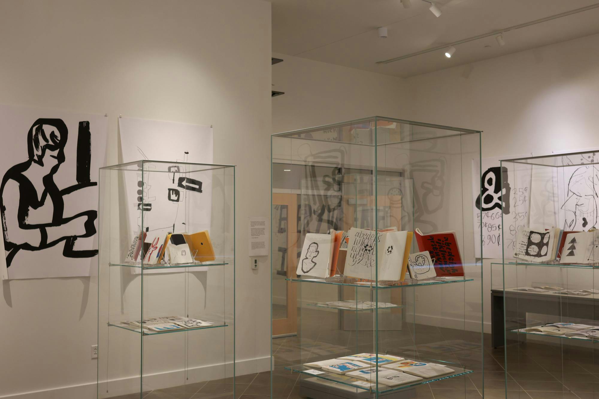 Photo of the “Everywhere I Go,” exhibit showing various clear display cases of open sketchbooks and large, bold black-and-white drawings displayed on the walls.