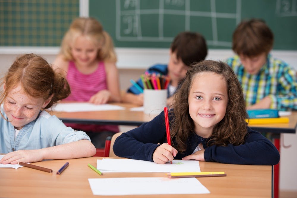 Beautiful happy young girl in class at school looking up from her desk and notes to smile at the camera