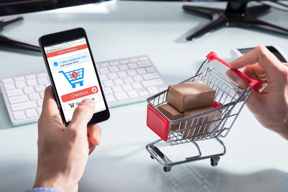 A Person's Hand Holding Cart With Cardboard Boxes While Shopping Online On Mobile Phone