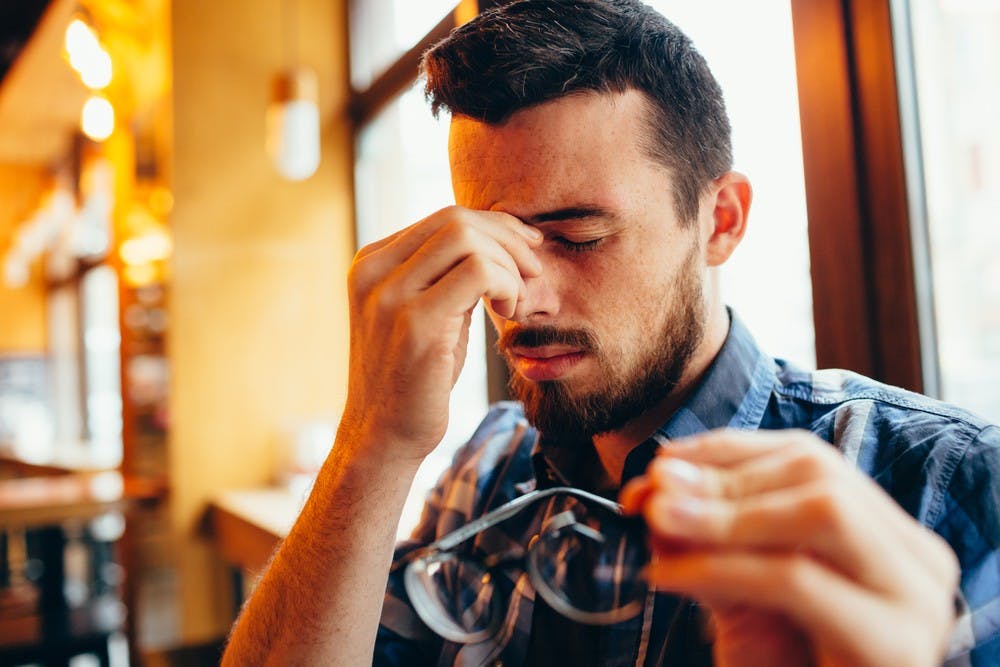 Closeup portrait of attractive man with glasses. Poor young guy has eyesight problems. He is rubbing his nose and eyes because of their weariness and holding his eyeglasses with one hand