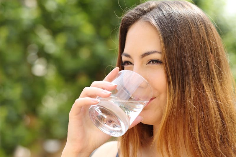 Happy healthy woman drinking fresh water from a glass outdoor with a green background            