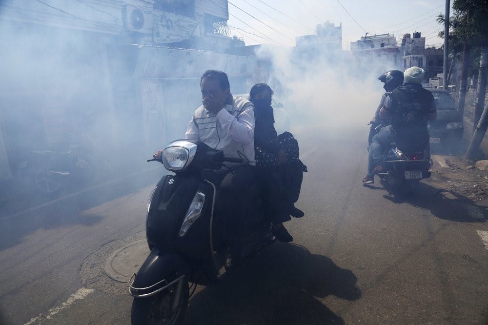 Indians ride two wheelers through smoke as municipal workers fumigate an area as a precautionary measure against the spread of new coronavirus March 16, 2020, in Jammu, India. For most people, the new coronavirus causes only mild or moderate symptoms. For some, it can cause more severe illness, especially in older adults and people with existing health problems. (AP Photo/Channi Anand)