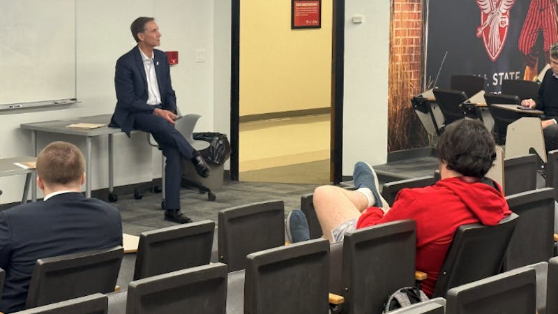 A man in a suit sits on a table speaking to an audience of students in a classroom setting.