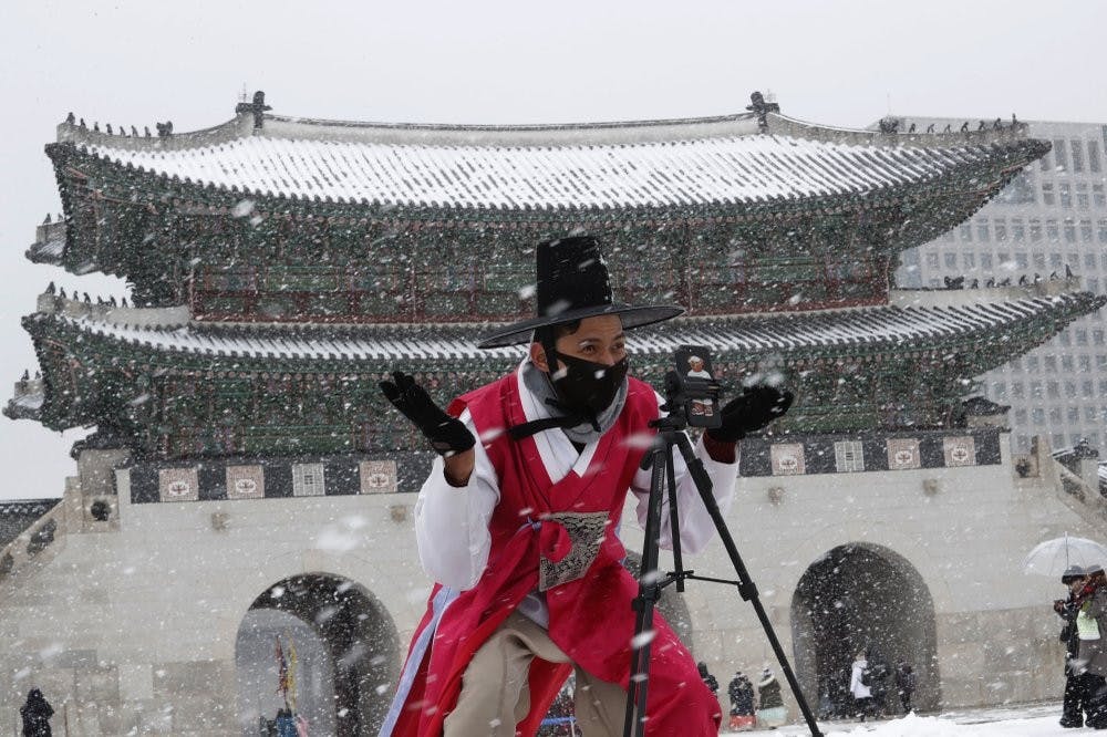 <p>A man dressed in South Korean traditional "Hanbok" attire wears a face mask in the snow as he gestures to take photos Feb. 17, 2020, at the Gyeongbok Palace, the main royal palace during the Joseon Dynasty in Seoul, South Korea. Chinese authorities on Monday reported a slight upturn in new virus cases and hundred more deaths for a total of thousands since the outbreak began two months ago. <strong>(AP Photo/Ahn Young-joon)</strong></p>