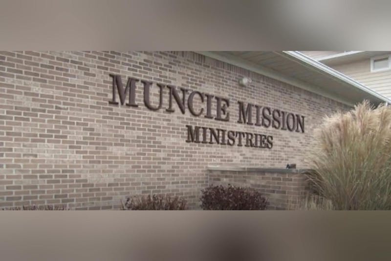 A brick wall features the words "Muncie Mission Ministries" in large, dark lettering, with some ornamental grass beside it.