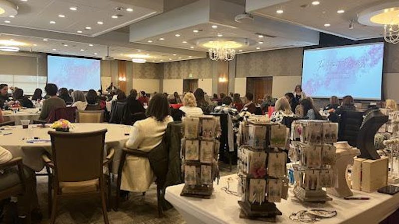 A spacious room with attendees seated at tables, focused on presentations displayed on large screens at the front.
