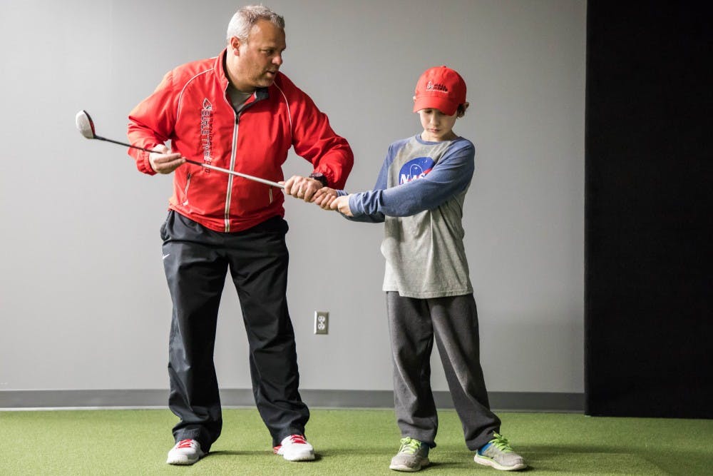 <p>Ball State men's golf head coach Mike Fleck works with Manasseh Crum on his swing Jan. 23 at the Travis Smith Short Game Center. Crum has Polyarticular Juvenile Idiopathic Arthritis. He is part of Team IMPACT, a national non-profit organization that connects children facing serious or chronic illnesses with college athletic teams. <strong>Rachel Ellis, DN</strong></p>