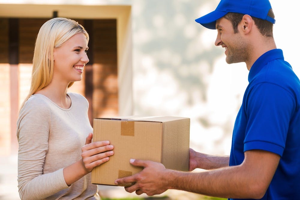It always arrives in time. Side view of happy young delivery man giving a cardboard box to young woman while standing in front of the house
