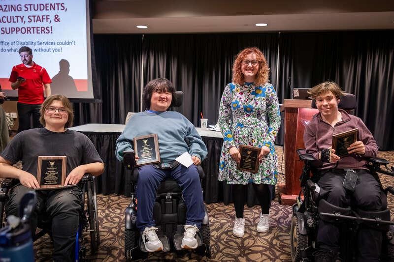 Four individuals in wheelchairs, holding plaques, stand in front of a presentation backdrop at a recognition event.