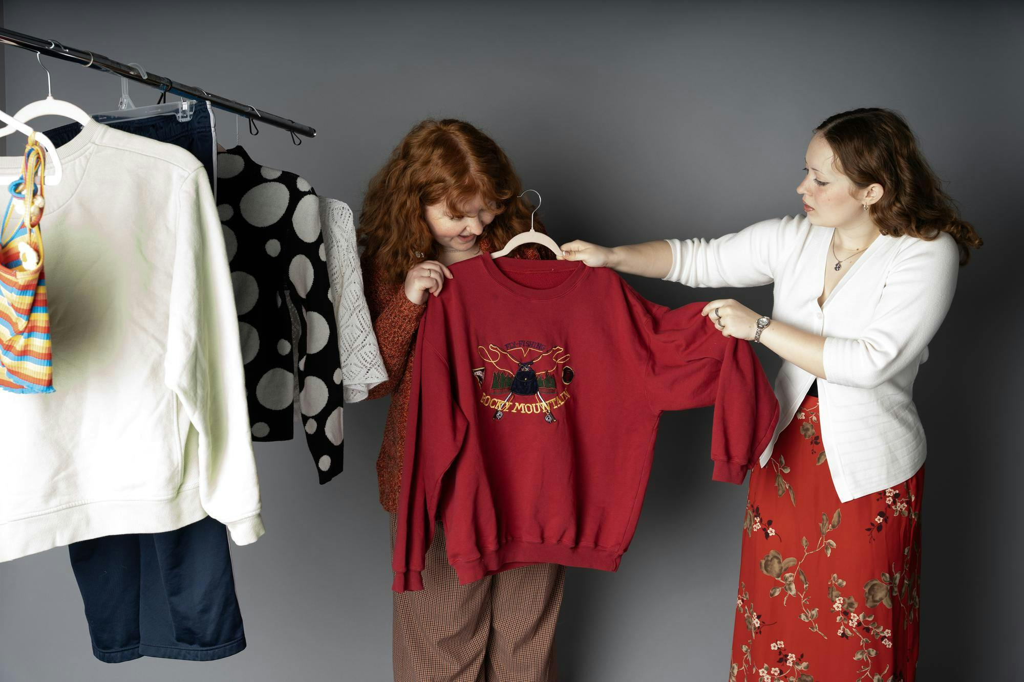 Two women pose in front of a clothing rack full of thrifted close. The brown haired woman, wearing a red floral dress and a white button down, holds up a red sweatshirt in front of a woman with red hair.