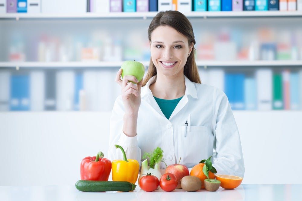 Smiling nutritionist in her office, she is holding a green apple and showing healthy vegetables and fruits, healthcare and diet concept