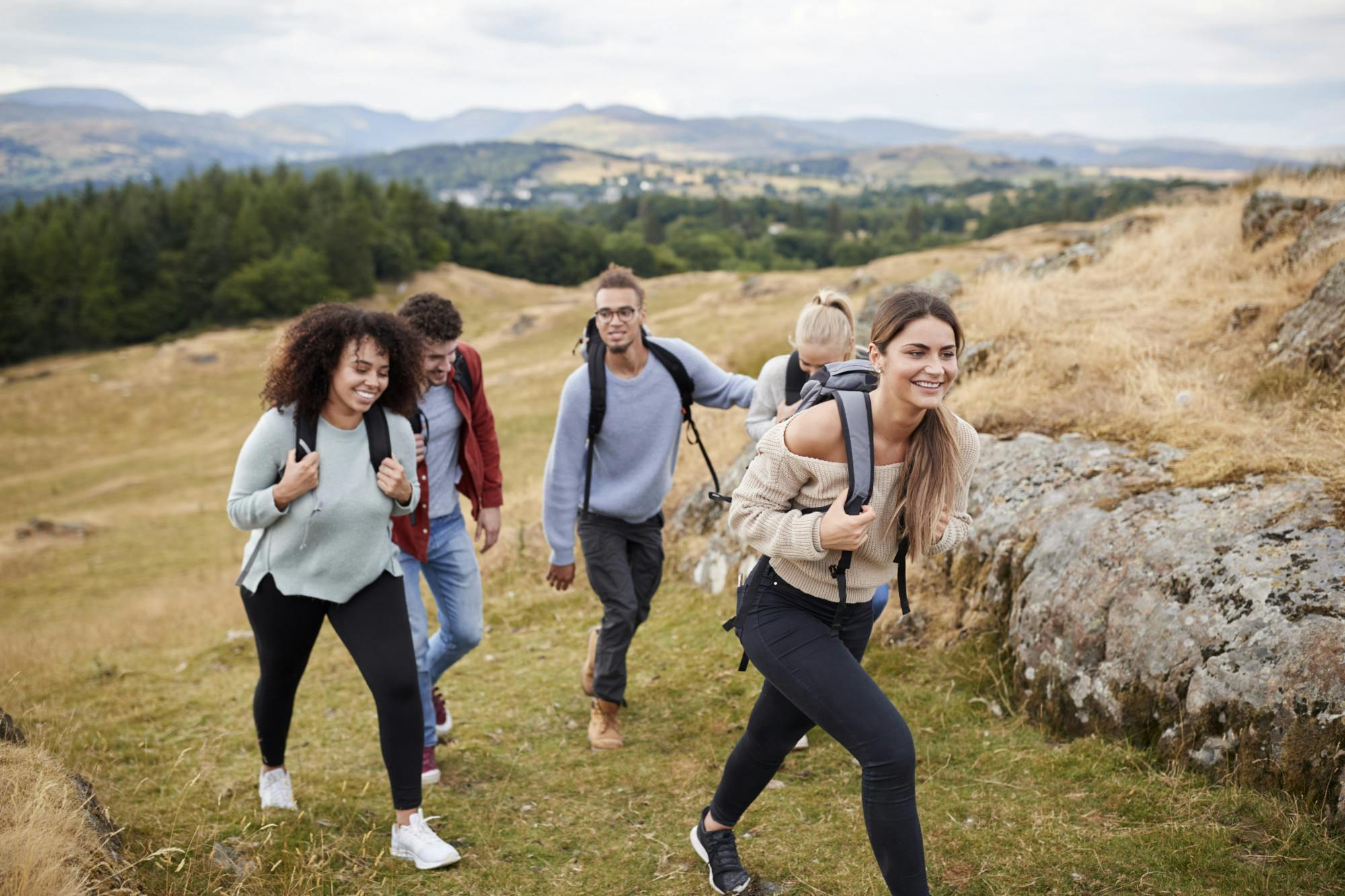 Multi ethnic group of five young adult friends hiking across a field uphill towards the summit, close up