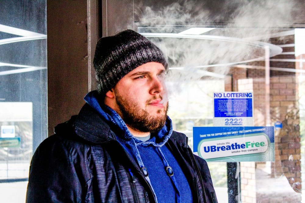 <p>A student smokes his vape outside Lockwood Library next to a smoke free sign. Vaping is prohibited under UB’s smoke-free policy. Students and faculty feel the university doesn’t do a good job enforcing its policy.</p>