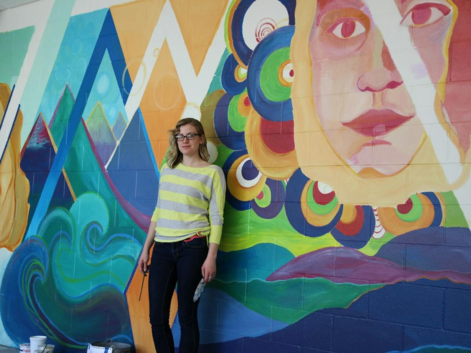 Natalie Miller, a senior fine arts major, poses by her near-completed final project for Thematic Drawing. Her mural on the CFA wall depicts two faces, one blue and one red, in an exploration of self-identity and the multiple sides of an individual’s inner reality.