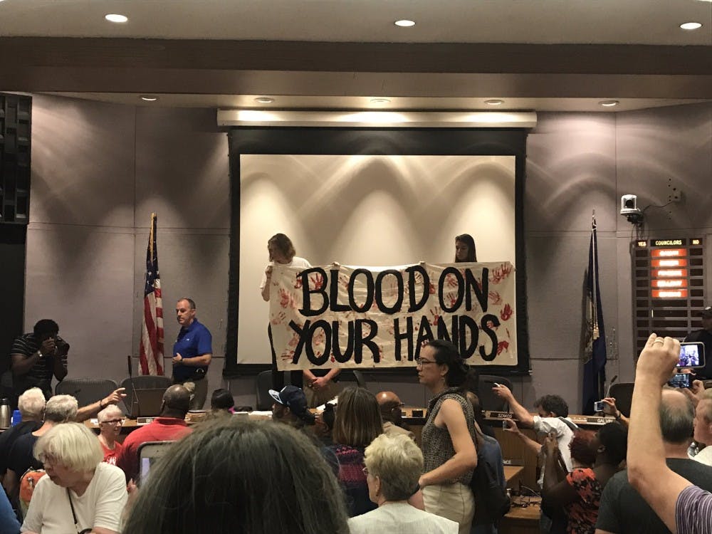 <p>Two audience members climbed atop the dais in front of City Council members holding a banner reading “Blood on your hands,” resulting in the departure of the councilors under police protection. &nbsp;</p>
