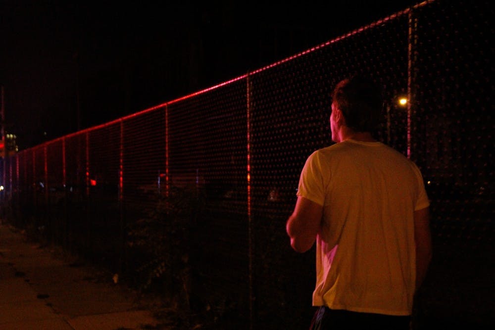 A resident smoked a cigarette while walking along the gates to the old West Philadelphia High School. Police lights painted the fence red and blue. Police officers directed all residents and bystanders away from the area on 48th and Walnut streets.