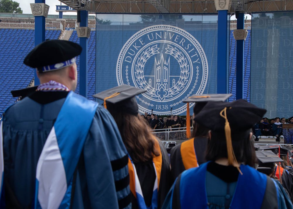 <p>Duke University logo framed by individuals participating in Commencement ceremony, in ceremonial robes, with their backs turned to the camera.</p>