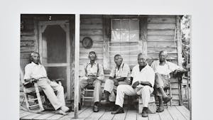 Black and white photograph from the Roland L Freeman Collection in Wilson Library. The photograph shows a group of 5 older black men sitting in wooden chairs on the front wooden porch of a wooden house. 
Photo Courtesy of Roland L. Freeman, Community Elders, Mississippi, July 1975.