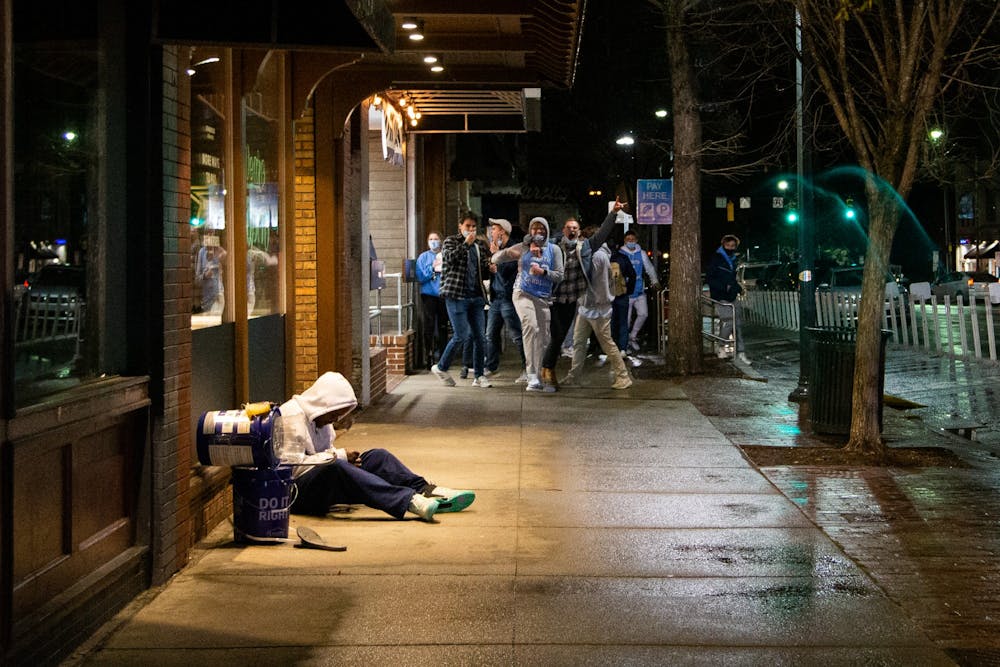 Students celebrating UNC's basketball team triumphs over Duke prepare to rush Franklin Street on Saturday, Feb. 6, 2021, while an unhoused man sits under an awning to stay out of the rain.