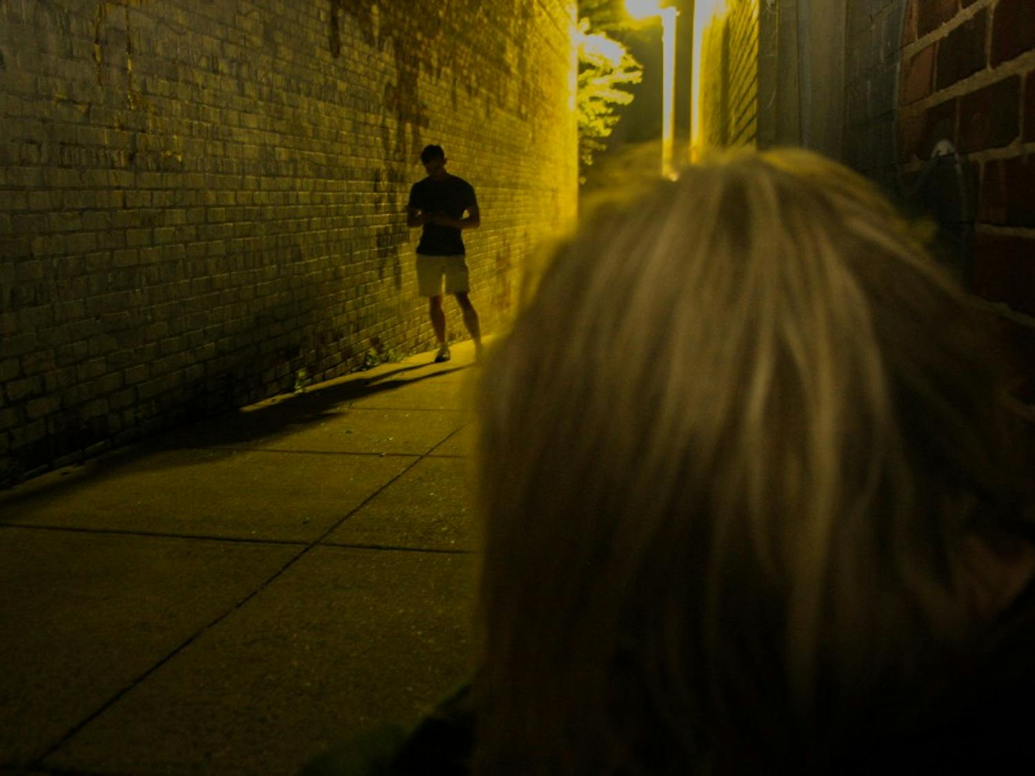 DTH Photo Illustration. A woman sits in an alley, alone, while a man on his phone walks by paying no attention to her.