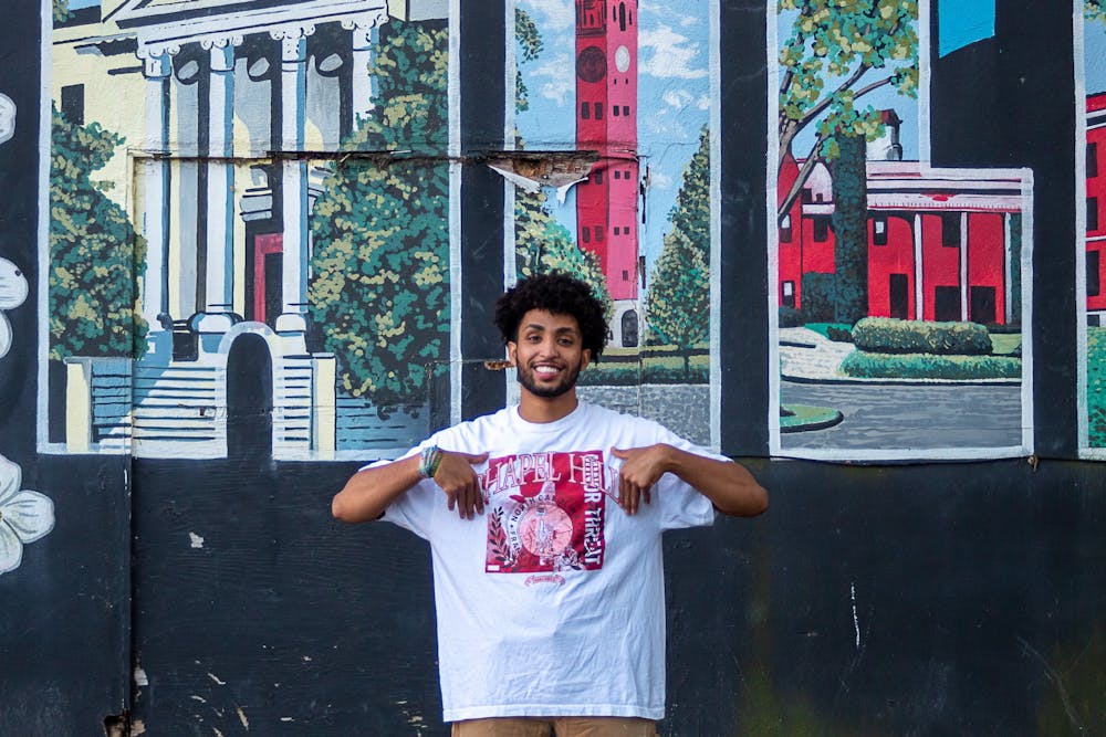 <p>Eliam Mussie, the co-president of the Black Entrepreneur Initiative, pictured in front of the "Greetings from Chapel Hill" mural on Rosemary Street in Chapel Hill, N.C. on Tuesday, Sept. 6, 2022. Mussie is wearing a shirt from his clothing brand, Franklin Street Market, which will be represented at the pop-up this coming Saturday, Sept. 10, 2022.</p>