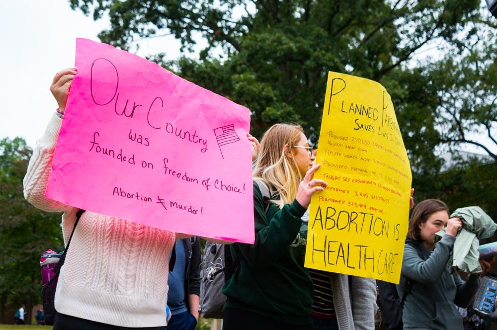<p>Students Erin Campagna and Braidyn Davis hide faces from the anti-abortion demonstrators with the Genocide Awareness Project that are attempting to photograph the students protesting the exhibit at Polk Place on Tuesday, Oct. 22, 2019. This exhibit included graphic images and made comparisons between abortion procedures and mass genocides.</p>