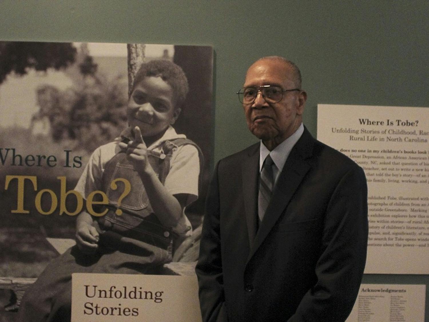 Charles Garner stands next to the book cover of "Tobe," which features a photograph taken of him as a child.   This photo is hanging in the North Carolina Collections Gallery, of Wilson Library.