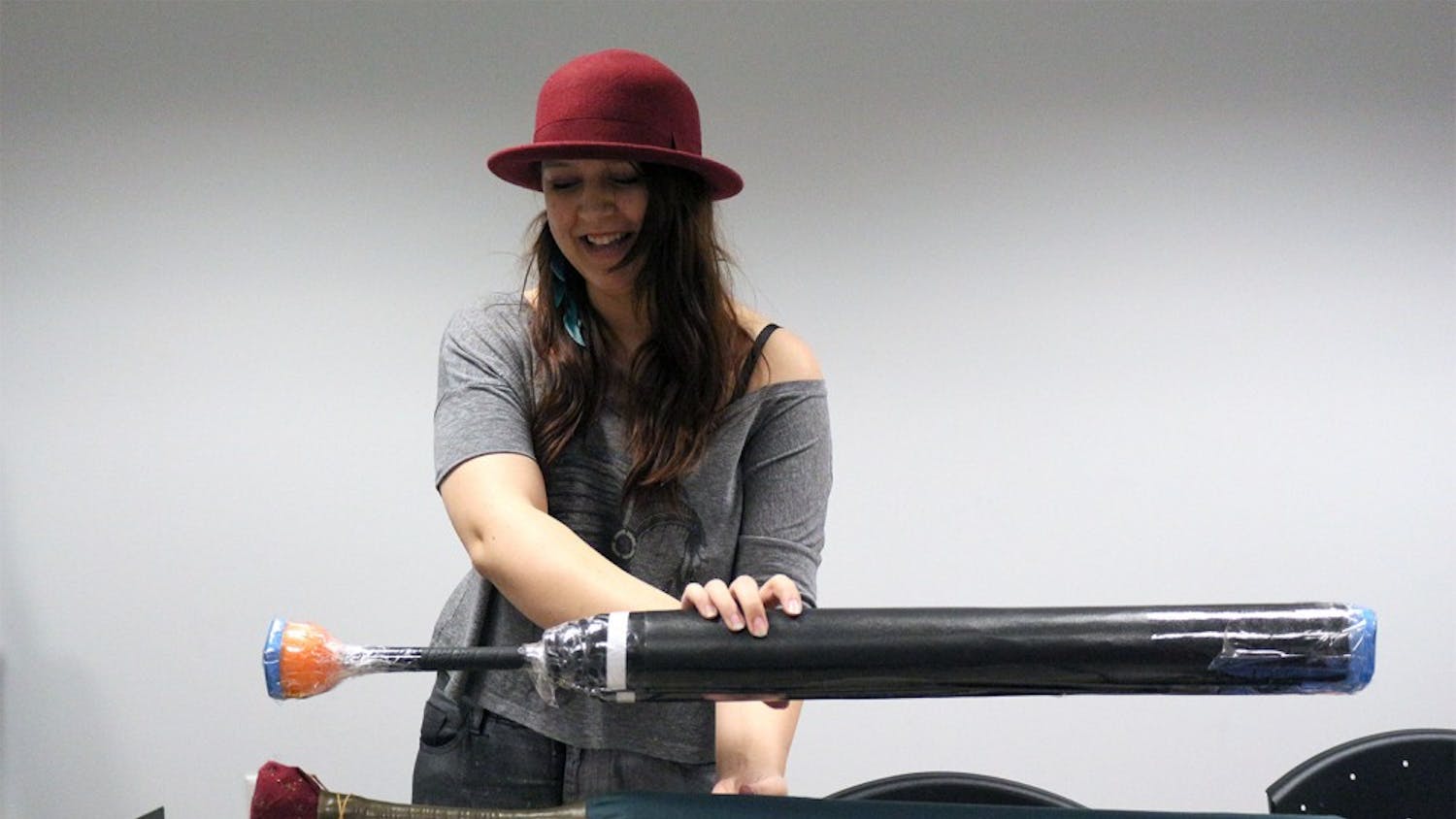 Alex Ruba holds up two different versions of a short sword, a type of weaponry used in game play. The top one is in the process of being made and shows the inner parts of the sword including the golf shaft center, the foam body, and the cut pool noodle at the top. The bottom sword is completed with a fabric covering. 